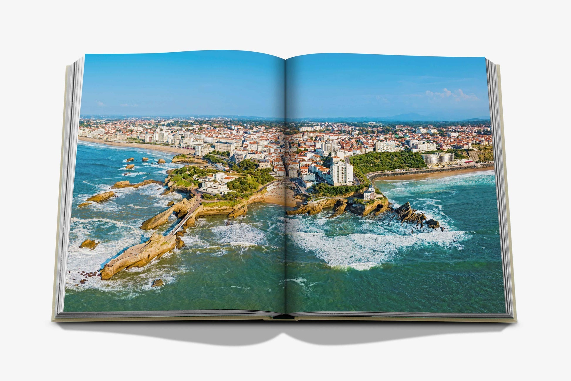 **Product Name: Biarritz Basque** by **Assouline** showcases an aerial photograph of Biarritz, a coastal city in the Basque Country. The image captures waves crashing against rocky shores, with buildings in the background—a paradise for surfing enthusiasts.