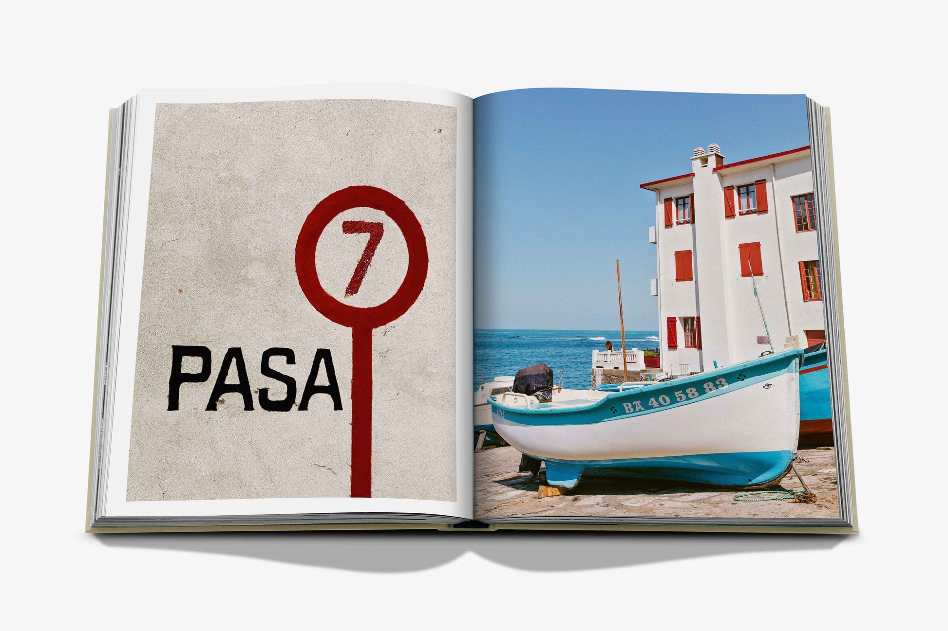 Open book showing two pages: the left page displays the text "PASA 7" with a red and black circular sign, while the right page features a white fishing boat near a seaside building with red shutters in the picturesque Basque Country. This image is from Biarritz Basque by Assouline.