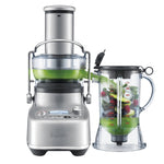 The Breville Bluicer Pro, a silver blender juicer with Cold Spin Technology, pours green juice into a jug of cut fruits and veggies, all shown on a white background.