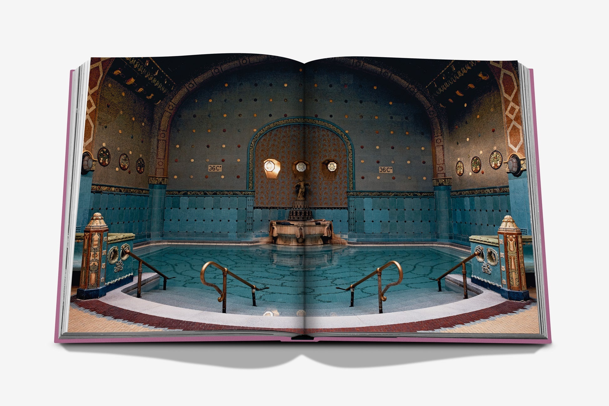 An open book, titled "Budapest Gem" by Assouline, reveals an image of an ornate indoor swimming pool reminiscent of Budapest's grandeur, adorned with mosaic tiles, columns, and decorative walls.