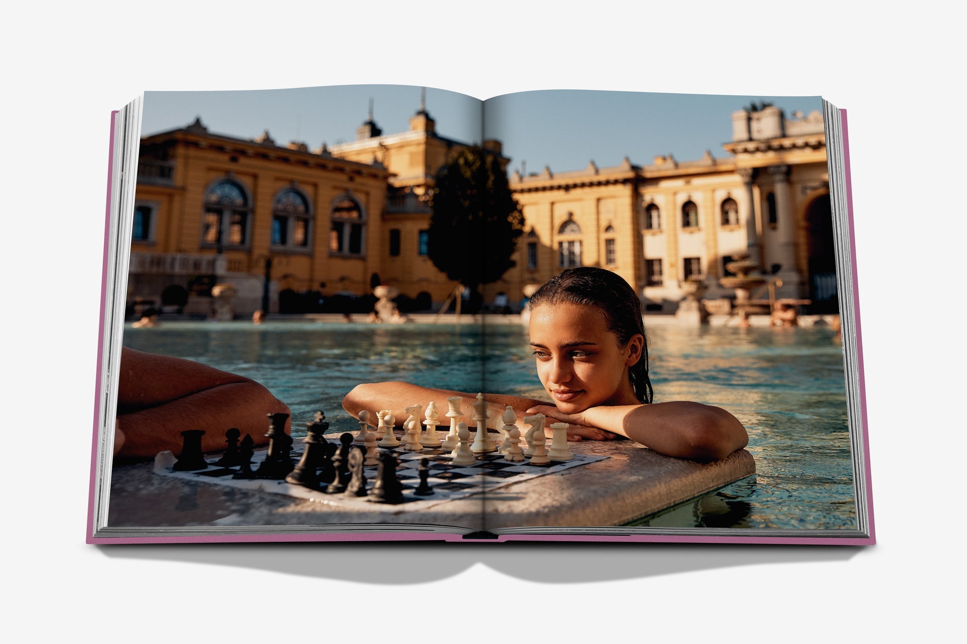 A person in a pool gazes intently at a chessboard on a floating platform, with the stunning Buda Castle visible in the background, embodying the allure of the Budapest Gem by Assouline.
