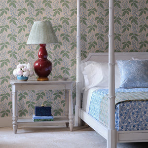 The Bunny Williams Keller Bed with patterned bedding and decorative pillows sits beside a bedside table topped with a red lamp, white flowers in a vase, and a book, all set against leafy wallpaper.