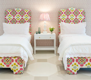 Two EJ Schrader Rosa Spring Twin XL Beds with matching colorful floral headboards and white bedding flank a white nightstand holding a lamp, books, and flowers, creating playful bedroom furniture ideal for any space.