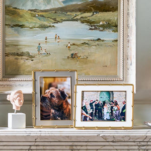 A framed landscape painting hangs above a marble mantel topped with two Addison Ross Gold and White Bamboo Frames (5x7) and a small bust, adding elegance with luxury velvet backs and gold-plated accents.