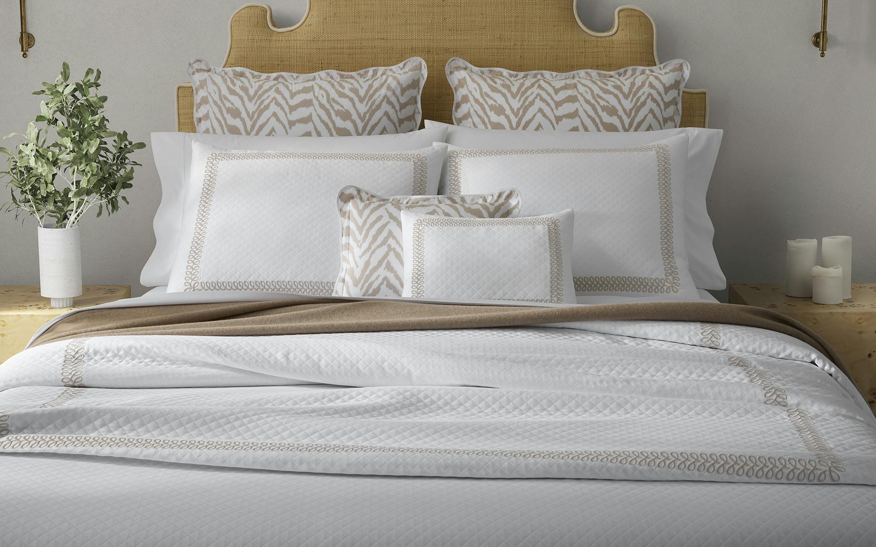 A neatly made bed with white and beige Astor Braid Matelassé Bedding Collection in Dune by Matouk, decorative pillows, a plant on the left nightstand, and three candles on the right nightstand.