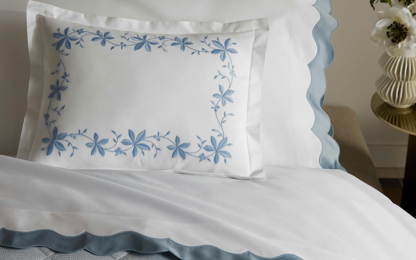 A white pillow with blue floral embroidery sits atop a neatly made bed dressed in Matouk’s Lorelei Bedding Collection, Hazy Blue.