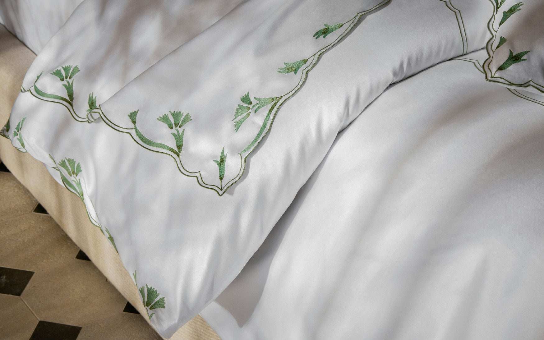Close-up of Matouk’s Marcella Bedding Collection in Clover, featuring green scalloped floral embroidery, draped elegantly over the side of a bed on a patterned floor.