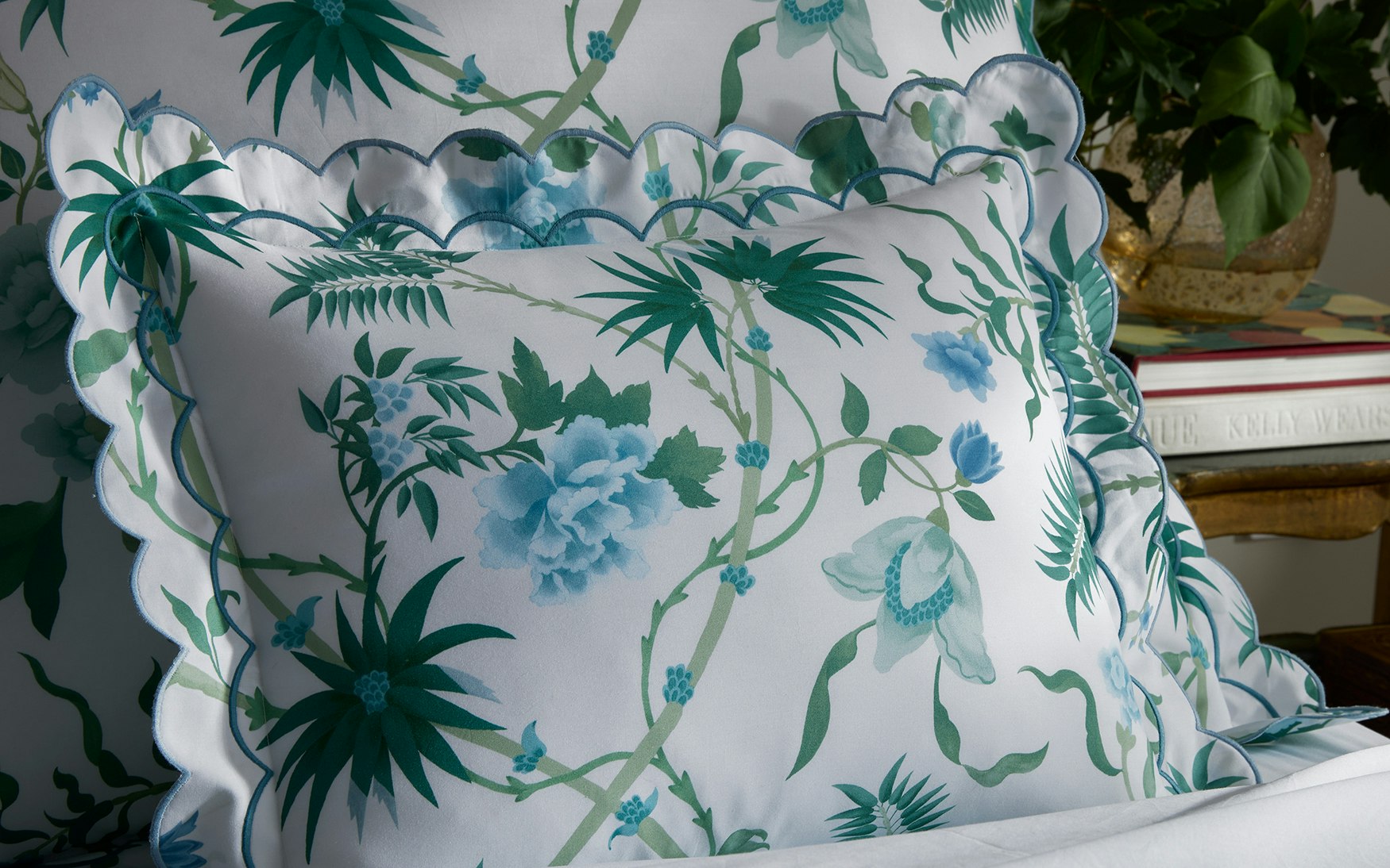 A decorative pillow with a scalloped edge and green-blue floral pattern rests on Matouk's Palmyra Bedding Collection in Emerald, with a stack of books and a potted plant in the background.