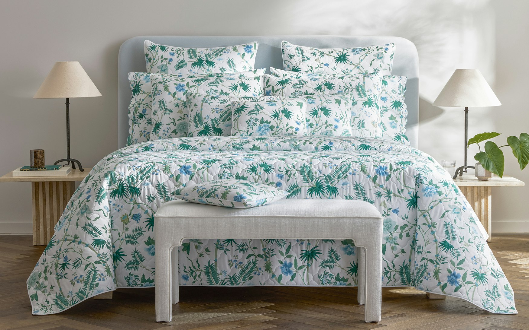 A neatly made bed features the Matouk Palmyra Bedding Collection in Emerald, accented by white and green palm fronds. Two lamps rest on side tables, with a bench positioned at the foot of the bed.