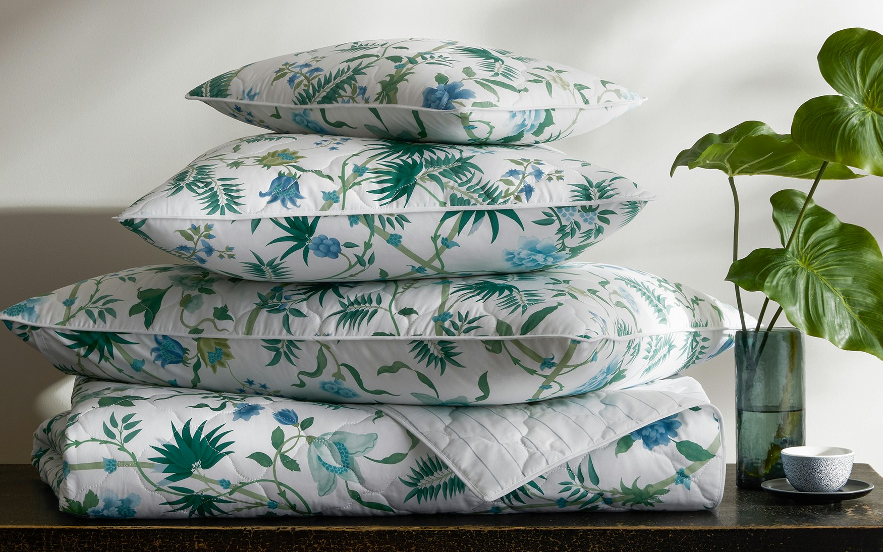 Four items from the Matouk Palmyra Bedding Collection in Emerald, featuring a green and blue tropical design, are stacked on a table beside a glass vase with green leaves and a small white cup.