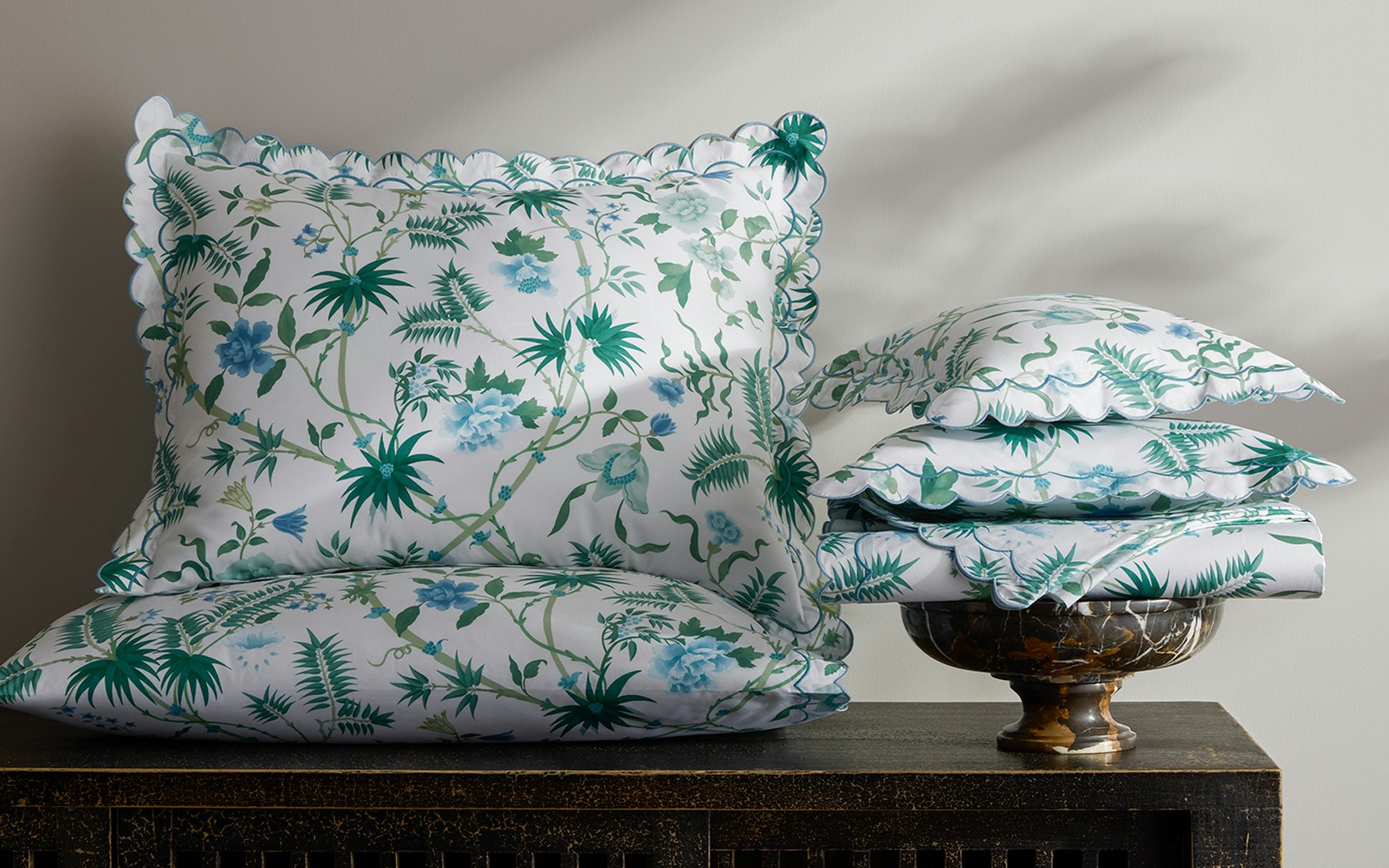 Four decorative pillows from Matouk's Palmyra Bedding Collection in Emerald, featuring blue and green botanical patterns with scalloped edges, are displayed on dark wood; three are stacked in a glass bowl.