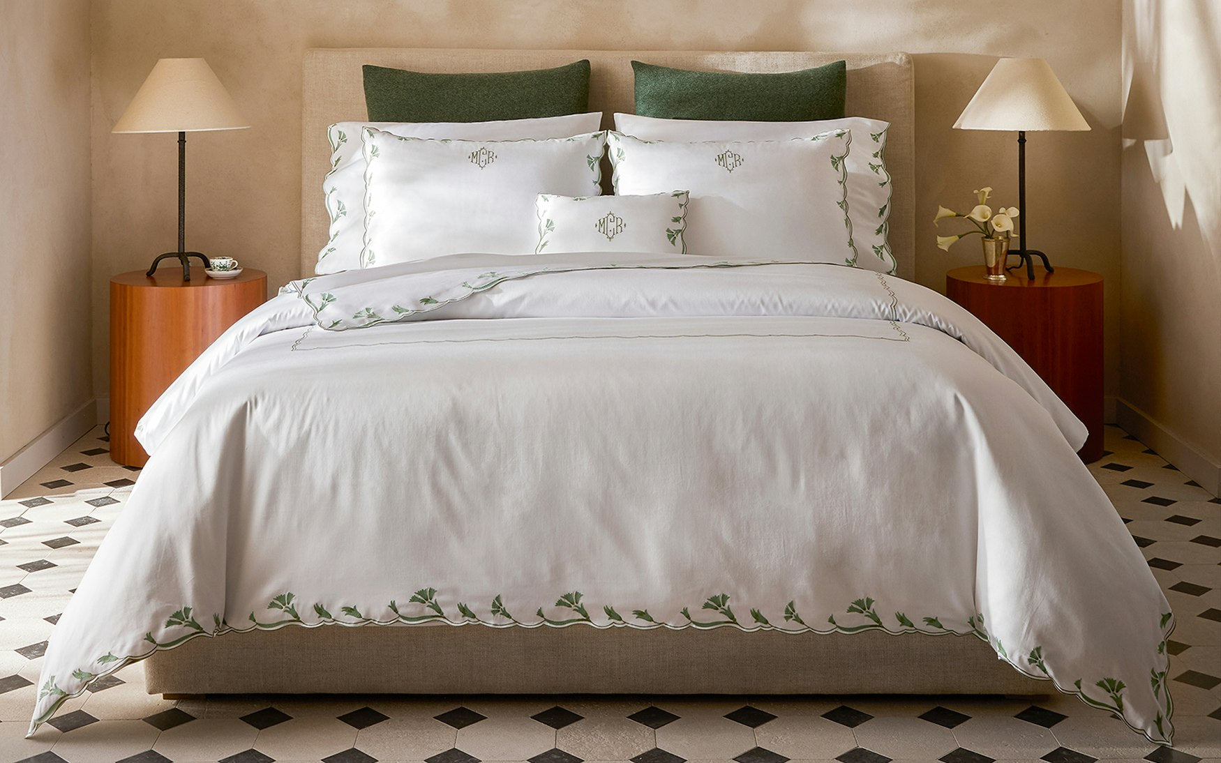 A neatly made bed features the Matouk Marcella Bedding Collection in Clover—white embroidered bedding with scalloped florals—three pillows, and two bedside tables with lamps, all set in a room with patterned floor tiles.