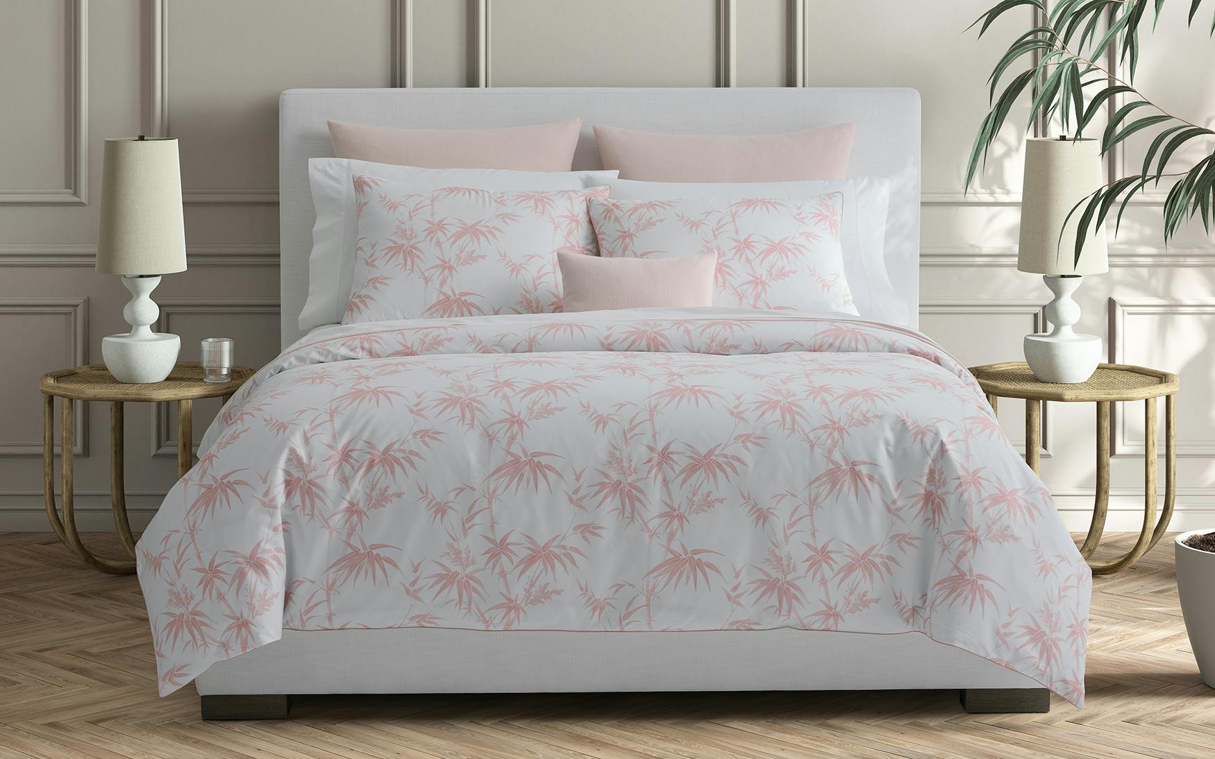 A neatly made bed with Matouk's Dominique Bedding Collection in Blush, crafted from OEKO-TEX® certified Egyptian cotton percale, flanked by two side tables with lamps, a plant, and neutral-colored walls in the background.