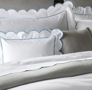 Neatly made bed with Matouk's Butterfield Bedding Collection in Ocean, featuring white pillowcases with blue trim, 500 thread count white sheets, and textured gray accent pillows and blanket.
