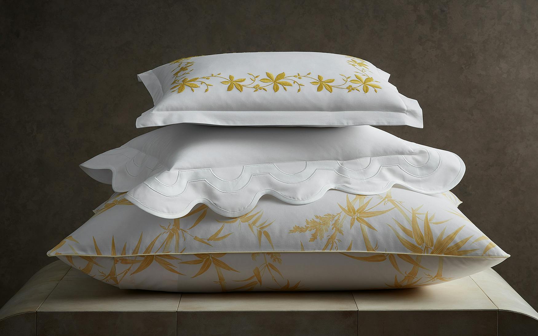 The Matouk Mirasol Bedding Collection in Bone features three stacked pillows with floral and decorative white and yellow patterns, made from luxurious Egyptian cotton percale with a 600 thread count, dramatically displayed against a dark background.