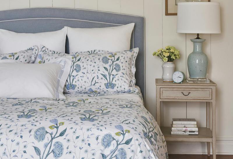 A bed is made up with Matouk's Schumacher Khilana Bedding Collection in blue, featuring a blue and white floral comforter, white pillows, and a gray headboard beside a nightstand topped with a lamp, clock, flowers, and stacked books.