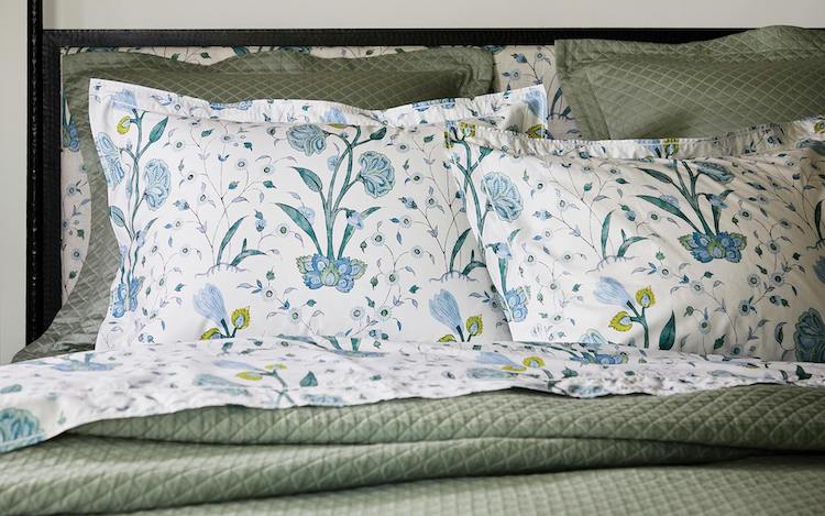 A bed featuring the Schumacher Khilana Bedding Collection in Blue by Matouk, with green quilted bedding and floral-patterned pillowcases and sheets in blue, green, and yellow hand-block textiles.