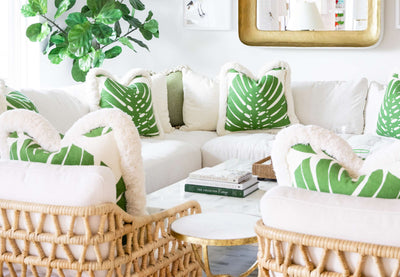 living room with white sofa, green and white pillows, fig leaf tree, gold mirror, and 2 woven cane chairs
