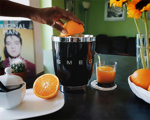 A hand presses a halved orange onto a Smeg Citrus Juicer Collection electric juicer on the kitchen counter, surrounded by a glass of juice, sliced orange, sunflowers, and a sugar bowl.