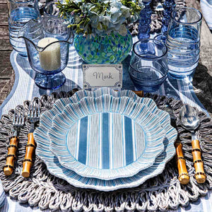 A table setting featuring Juliska’s Cabana Stripe Melamine Dinnerware Collection, bamboo-handled cutlery, blue glassware, a candle, a floral centerpiece, and a place card labeled "Mark.