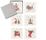 The Kim Seybert Holly Jolly Cocktail Napkin Set includes six square white linen napkins, each with a unique embroidered Christmas motif, shown beside a gray Kim Seybert box.