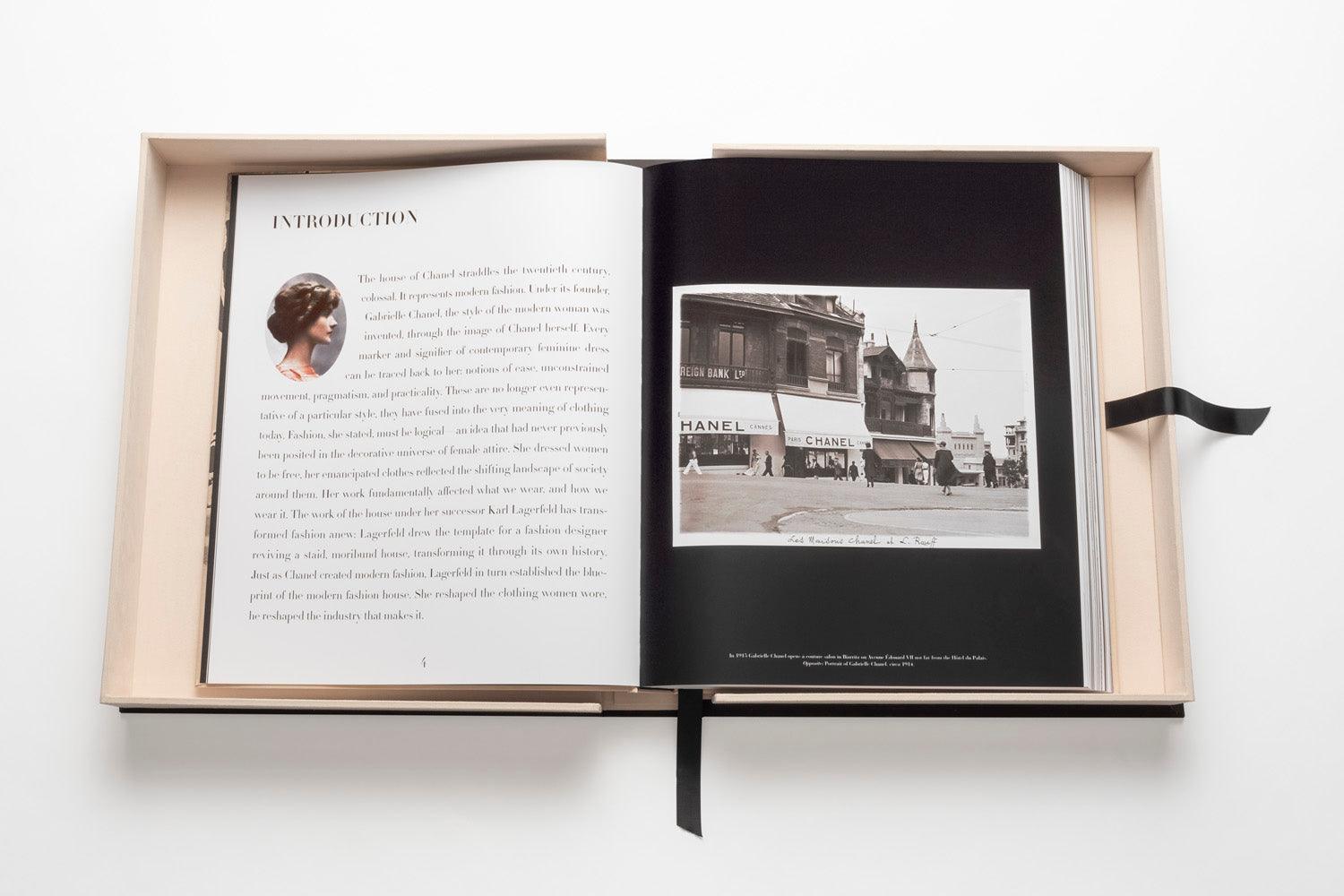 A large open book lies on a white surface, displaying text and a black-and-white photo of a Chanel storefront on the right page. The Impossible Collection of Chanel by Assouline, capturing Gabrielle Chanel's legacy, is nestled in a beige box with a black ribbon bookmark visible, exuding an air of luxe minimalism.