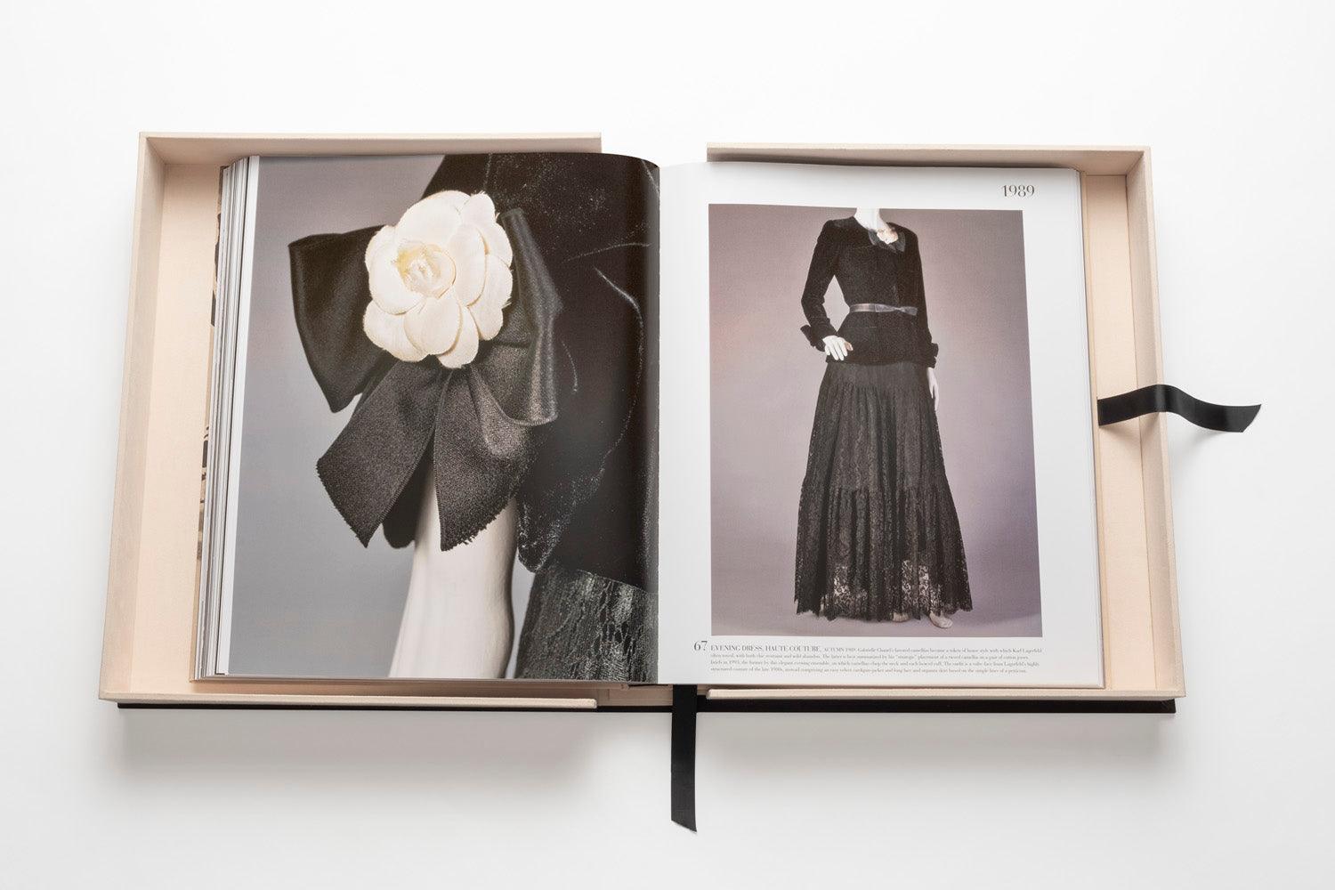 Open book showcasing fashion photographs. Left page features a close-up of a black outfit with a white flower accessory; right page shows a full-length black couture outfit with text above it, channeling luxe minimalism. The Impossible Collection of Chanel by Assouline placed in a light-colored box.