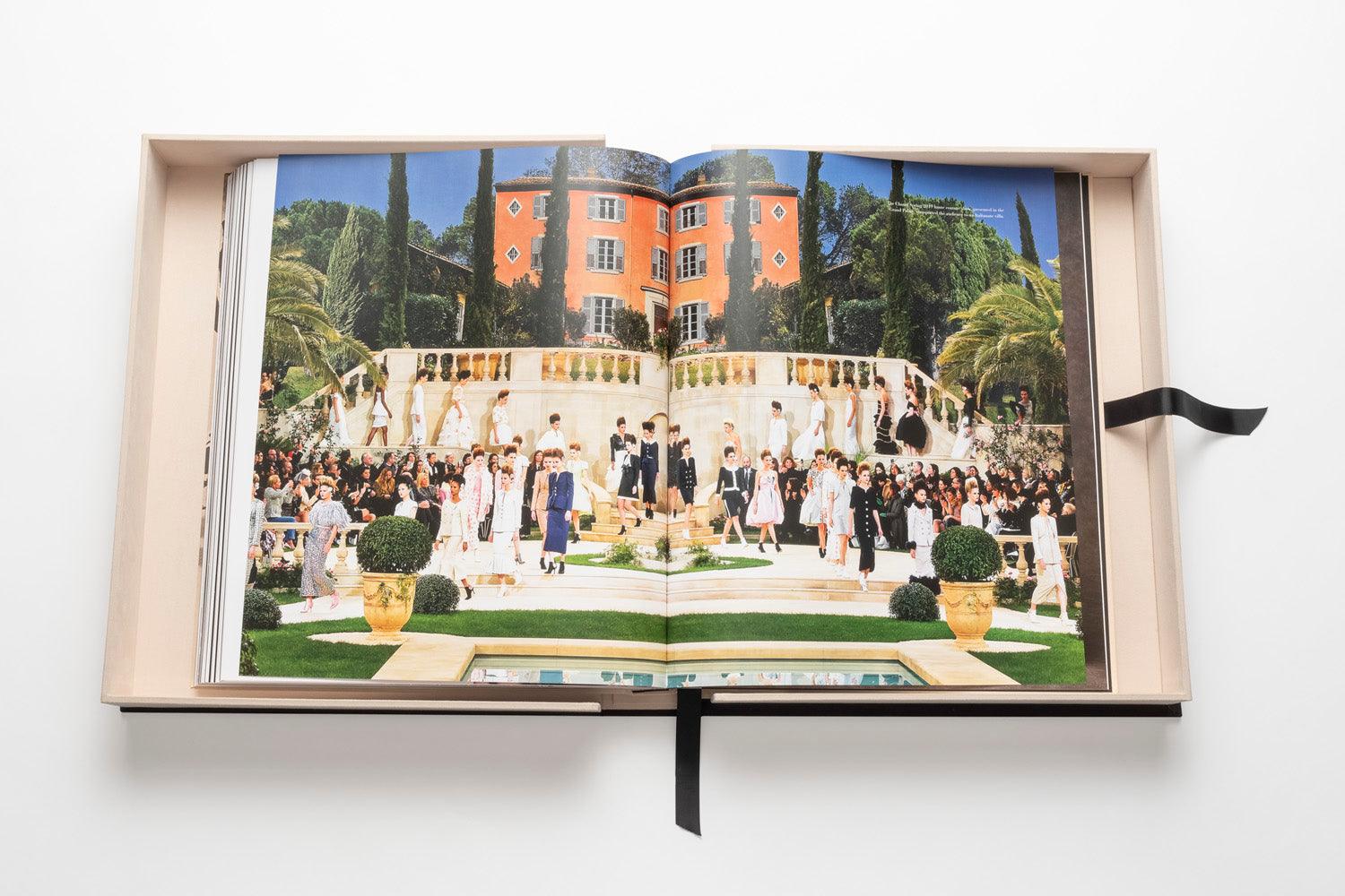 An open book displaying a colorful fashion runway show in front of a grand building with columns and a staircase, surrounded by palm trees and garden foliage, embodies understated luxury reminiscent of Gabrielle Chanel's timeless elegance. The book is titled "The Impossible Collection of Chanel" by Assouline.