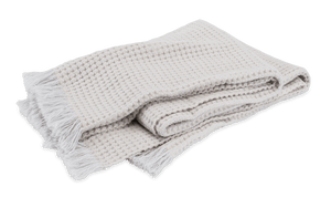 The Cleo Throw Collection by Matouk features a folded, light-colored cashmere throw with a subtle waffle-textured basketweave pattern and fringed edges, displayed on a plain background.
