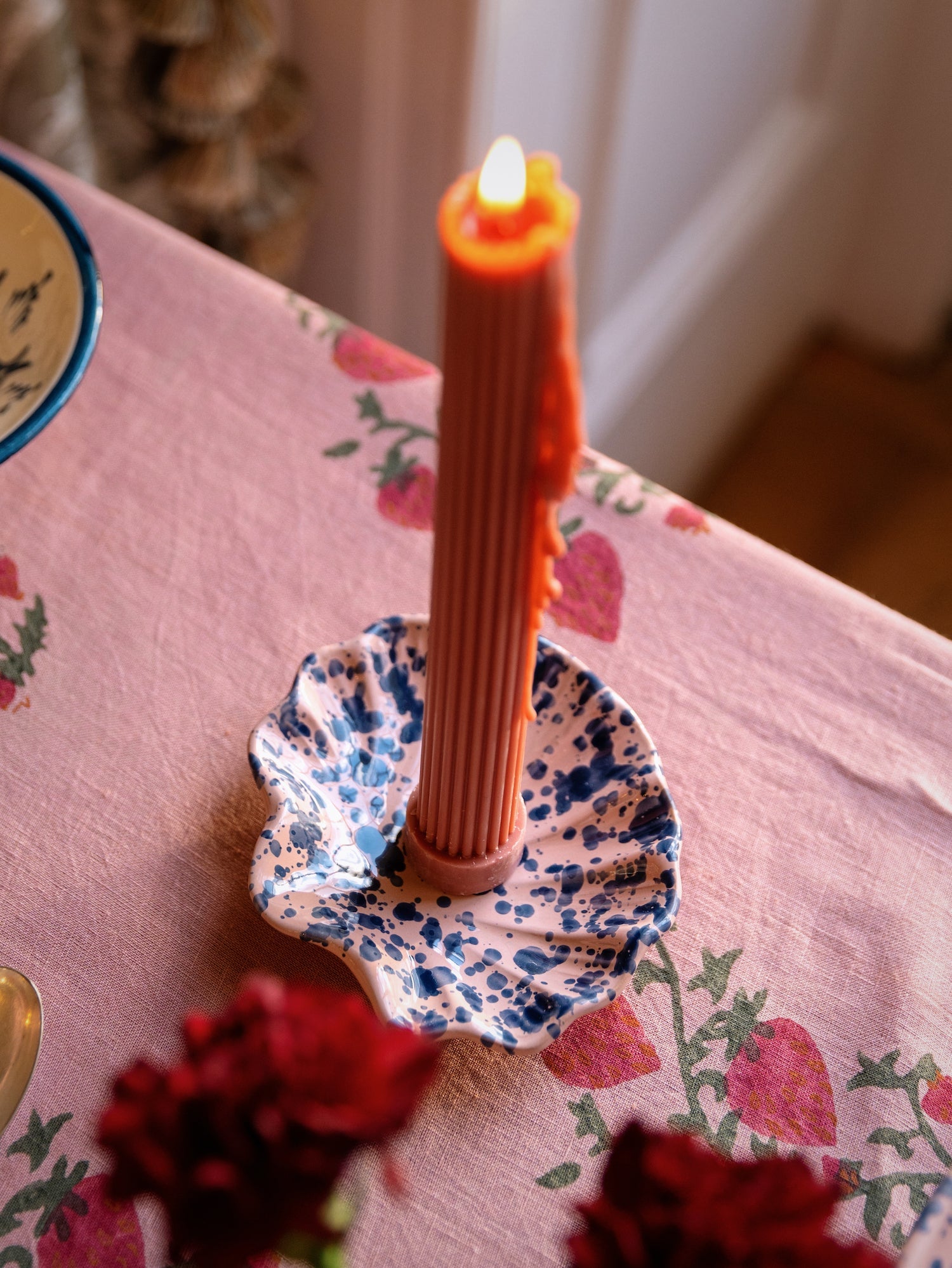 A ribbed orange candle in a Sharland England Splatter Coquillage Dish sits on a pink tablecloth with red floral patterns—an effortlessly chic, handmade-in-Italy gift.