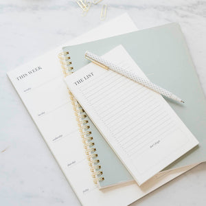 The Sugar Paper Cream List Pad, two notebooks, a white pen with black dots, and paper clips lie on a white marble surface, creating an elegant setup for organizing your lists.