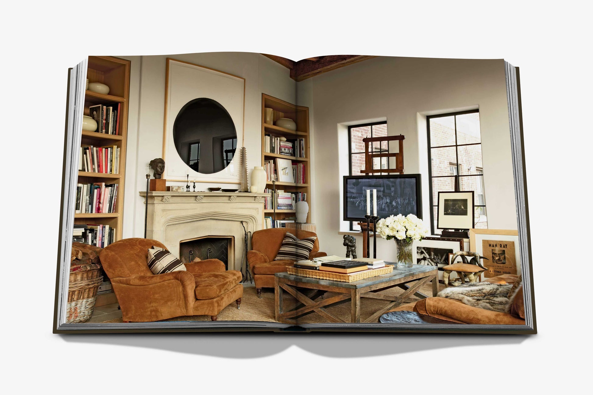 Assouline's "The Living Room" open book displays a cozy space with brown armchairs, fireplace, bookshelves, coffee table, and sunlit windows—offering an inviting look at modern minimalism in living room design.