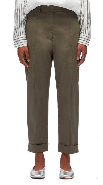 A person stands wearing 3.1 Phillip Lim olive green stretch cotton trousers with cuffed hems, paired with a white shirt featuring black stripes, and metallic silver shoes.
