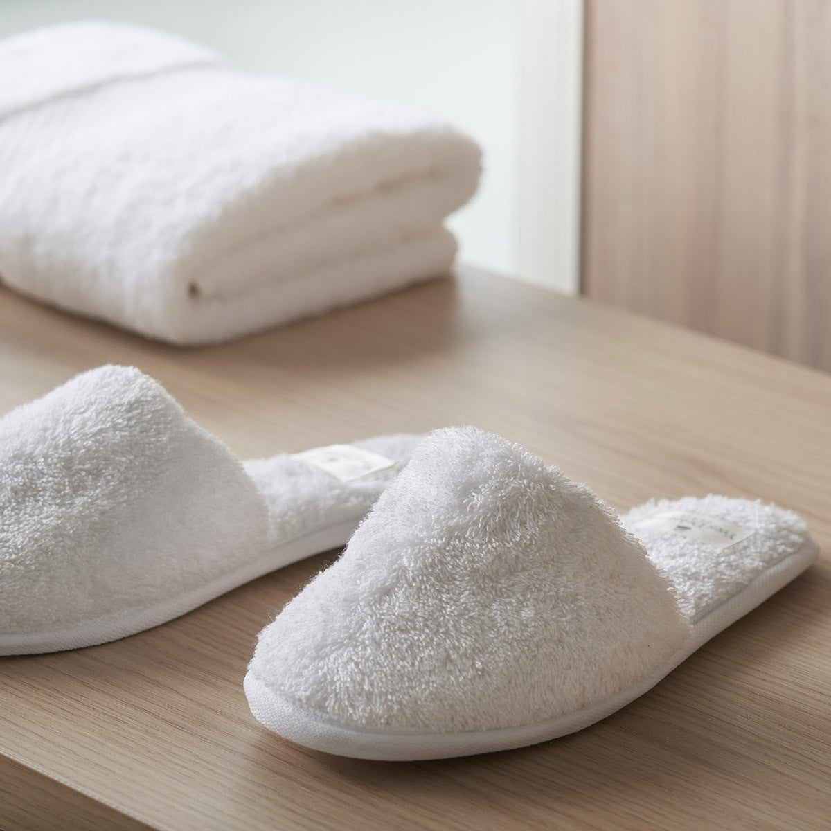 The Etoile Blanc Slippers by Yves Delorme, crafted from soft white terry cloth, rest on a wooden surface with folded absorbent long-staple cotton towels in the background for ultimate comfort.