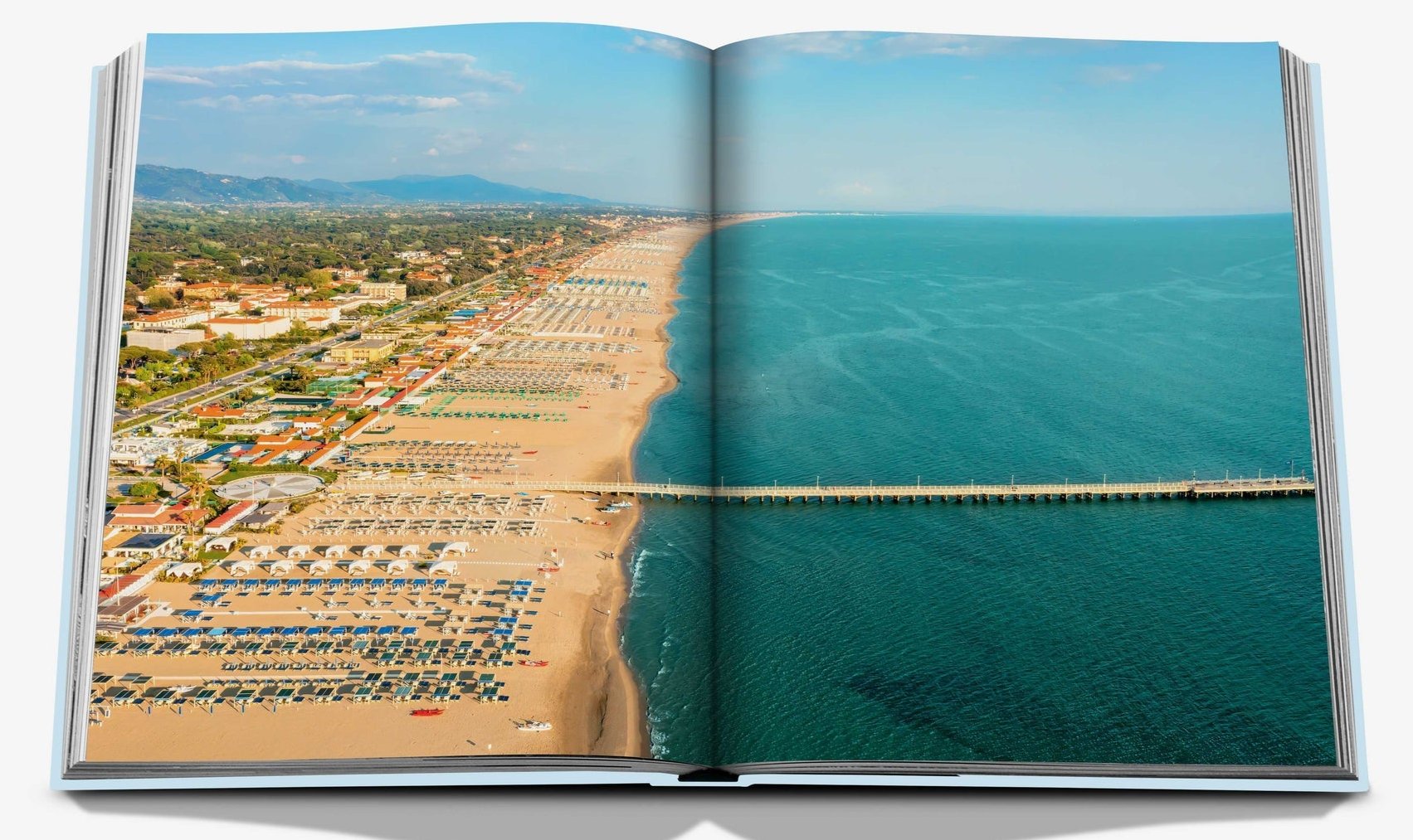 An open book from Assouline titled "Forte dei Marmi" displays a full-page aerial photograph of Tuscany's Mediterranean coast, showcasing a coastal beach with shoreline, rows of beach umbrellas, a pier, and adjacent green and urban landscapes.