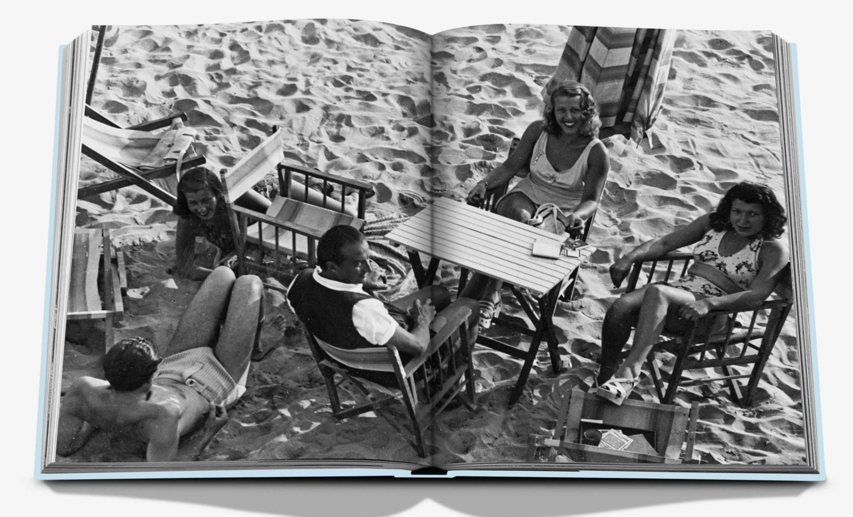 Black and white photo titled "Forte dei Marmi" by Assouline, capturing five individuals relaxing on a sandy beach in Forte dei Marmi. They are seated in chairs around a small table that holds a deck of cards and drinks.