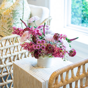 A Live Fall Floral Arrangement, Purple Ombre by Hive Floral Studio sits in a white vase on a striped table between two wicker chairs near a window.