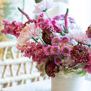 A white vase displays the Hive Floral Studio Live Fall Floral Arrangement, Purple Ombre, on a striped table with a wicker chair and patterned pillows in the background.
