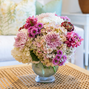The Hive Floral Studio Live Fall Floral Arrangement, Purple Traditional, featuring white, pink, and purple blooms in a glass vase, rests on a woven coffee table in a bright living room.