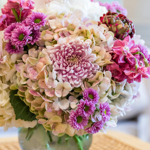 The Hive Floral Studio Live Fall Floral Arrangement, Purple Traditional, features a stunning mix of seasonal blooms artfully arranged and displayed on a light-colored woven surface.