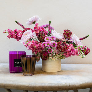 A Live Fall Floral Arrangement in a purple ombre vase with a candle from Hive Floral Studio sits on a table next to its packaging.