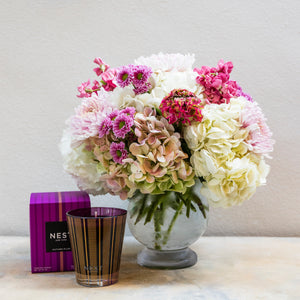 The Hive Floral Studio Live Fall Floral Arrangement, Purple Traditional with Candle, features seasonal pink and white flowers in a glass vase beside a purple-striped candle on a light surface.