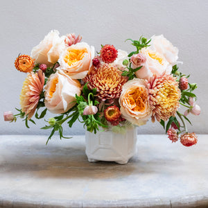 The Live Fall Floral Arrangement, Golden Ombre by Hive Floral Studio features peach roses, pink and orange chrysanthemums, and green foliage in a white ceramic vase on a light marble surface.