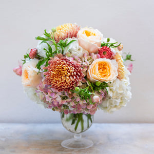 The Live Fall Floral Arrangement, Golden Traditional by Hive Floral Studio features peach, pink, and white roses, chrysanthemums, and hydrangeas in a glass vase—a lovely seasonal display set against a plain light background.