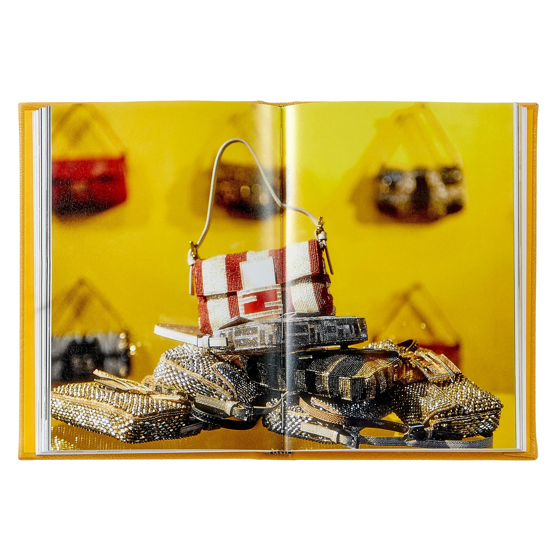 Open book, "Little Book of Fendi" by Graphic Image, open to a photo of luxury handbags—a red-and-white style perched on metallic clutches—set against a yellow backdrop, capturing the brand's iconic elegance.