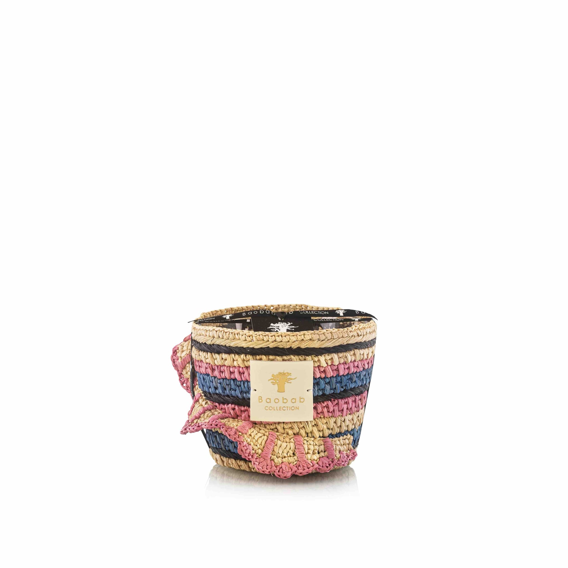 A woven basket with pink, blue, and natural horizontal stripes holds the "Baobab Collection Fety Mandihy Candle" by Baobab, making it a stunning decorative piece for any room.