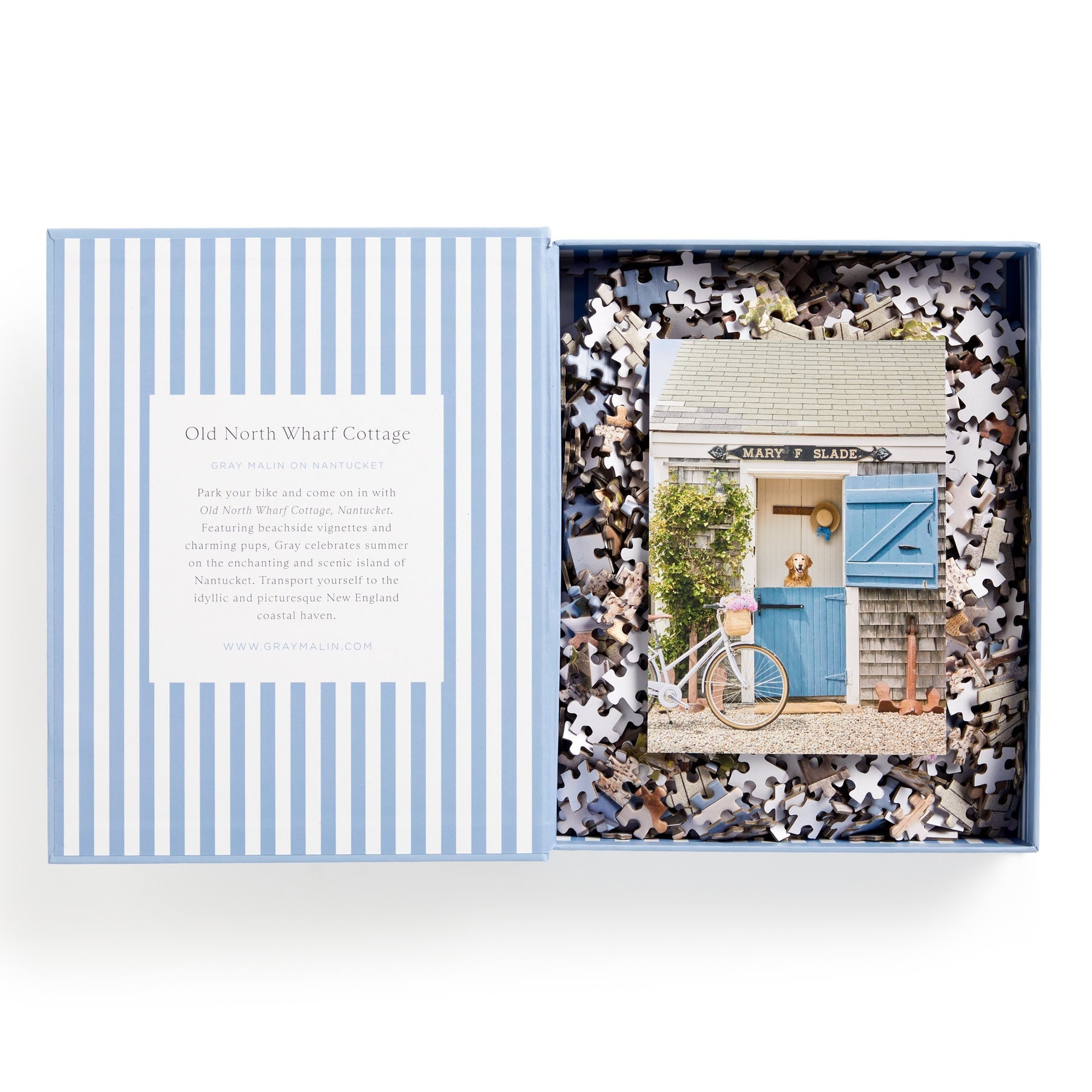 The Gray Malin 1000 Piece Puzzle Book Box, Nantucket by Chronicle Books unveils a charming cottage design with a light blue striped lid. Inside, discover a cozy cottage door, bicycle, and potted plant amid vibrant pieces capturing serene coastal scenes.