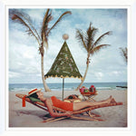 A woman in a red swimsuit unwinds on a beach lounger beneath a Christmas tree umbrella, echoing Slim Aarons' style in the iconic "Palm Beach Idyll," with palm trees and the ocean in the background. (Getty Images, Soicher Marin, January 1, 1955).