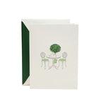 The Crane Garden Bistro Card Set features pearl white folded notes engraved with a bistro table, chairs, and a potted topiary, complemented by green-lined envelopes—perfect for sophisticated correspondence.
