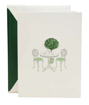 The Crane Garden Bistro Card Set features pearl white folded notes engraved with a bistro table, chairs, and a potted topiary, complemented by green-lined envelopes—perfect for sophisticated correspondence.